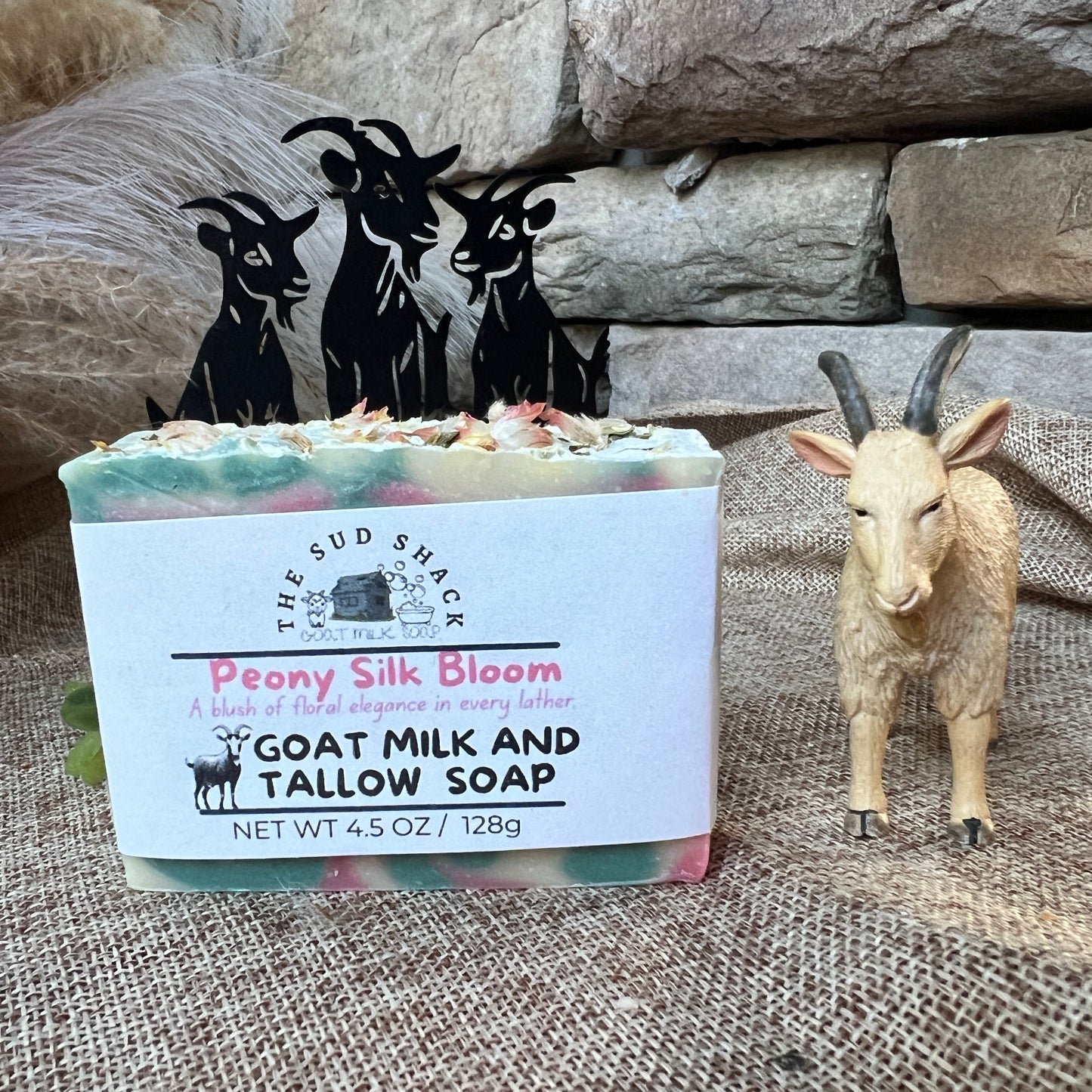 Bar of soap with a label featuring 'The Sud Shack' branding, surrounded by decorative goat figurines and a stone wall.