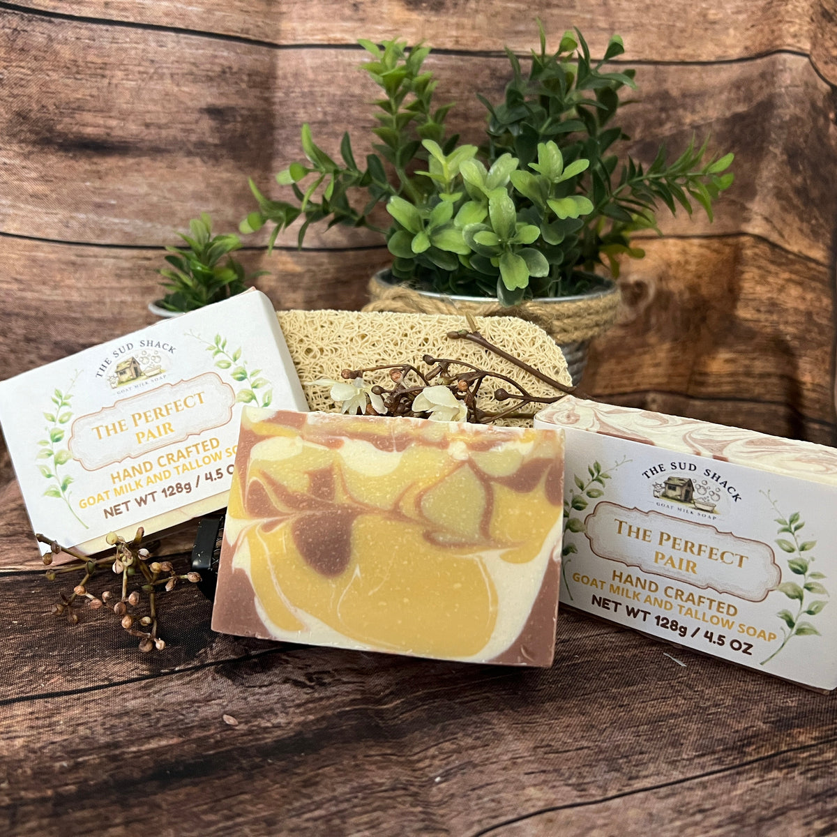 Three bars of handcrafted soap from The Kid's Nook, including 'The Perfect Pair' in Gold and Cream, displayed on a rustic wooden surface with a small potted green plant in the background.