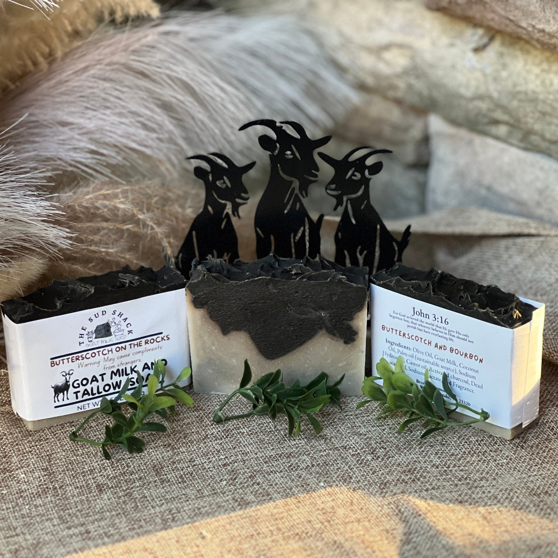 Three bars of soap with goat-themed packaging on a textured surface.