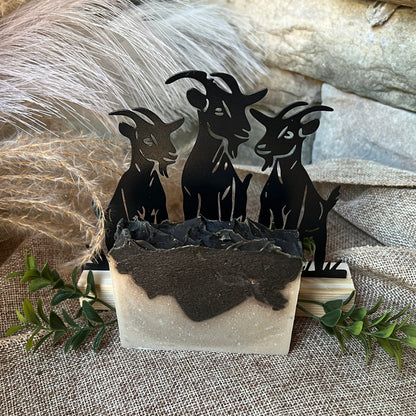Layered Soap with a Decorative set of black goat figures in a stone container on a textured surface with feathers and greenery.