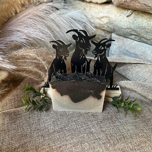 Layered Soap with a Decorative set of black goat figures in a stone container on a textured surface with feathers and greenery.