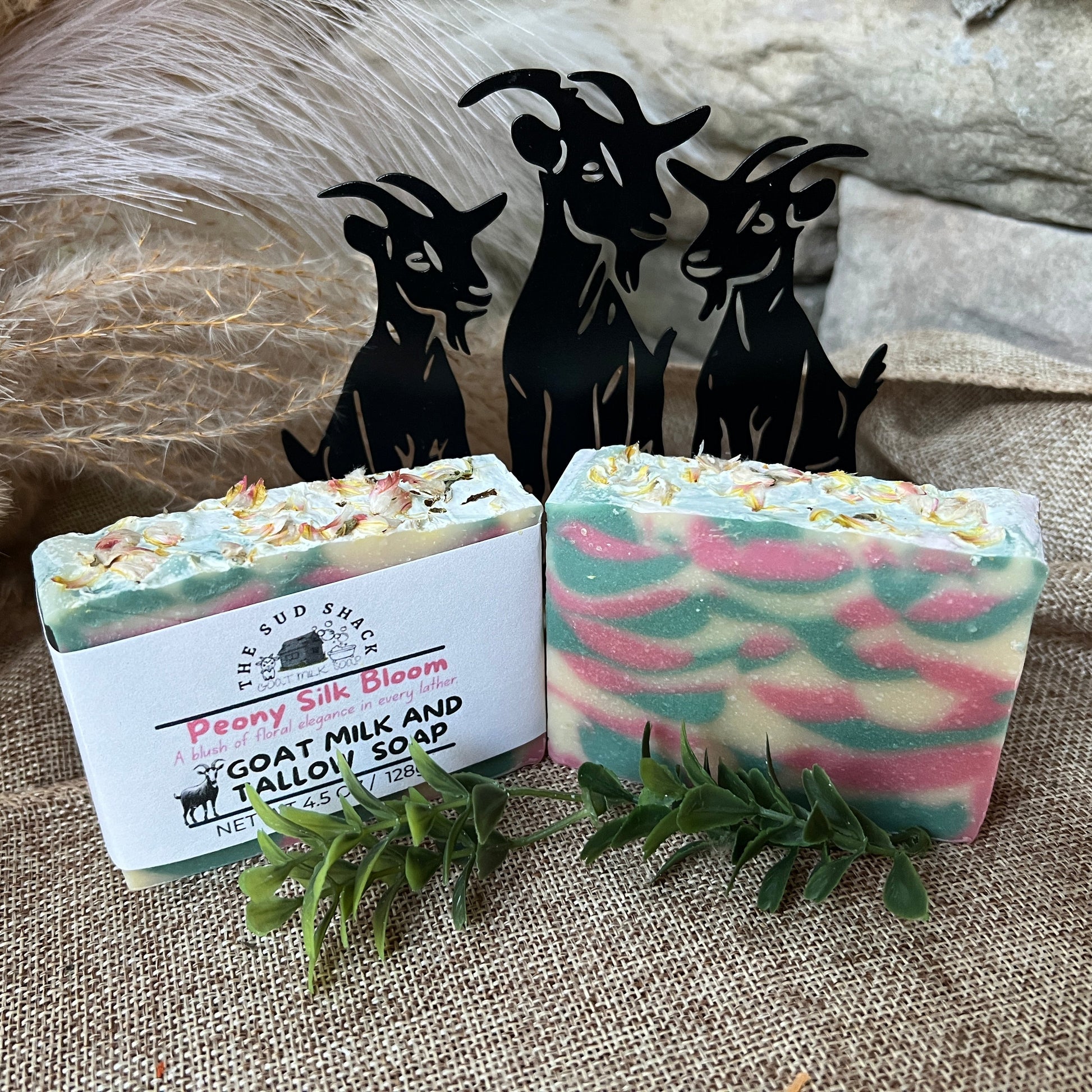 Two bars of soap with decorative packaging on a textured surface, accompanied by small goat figurines.