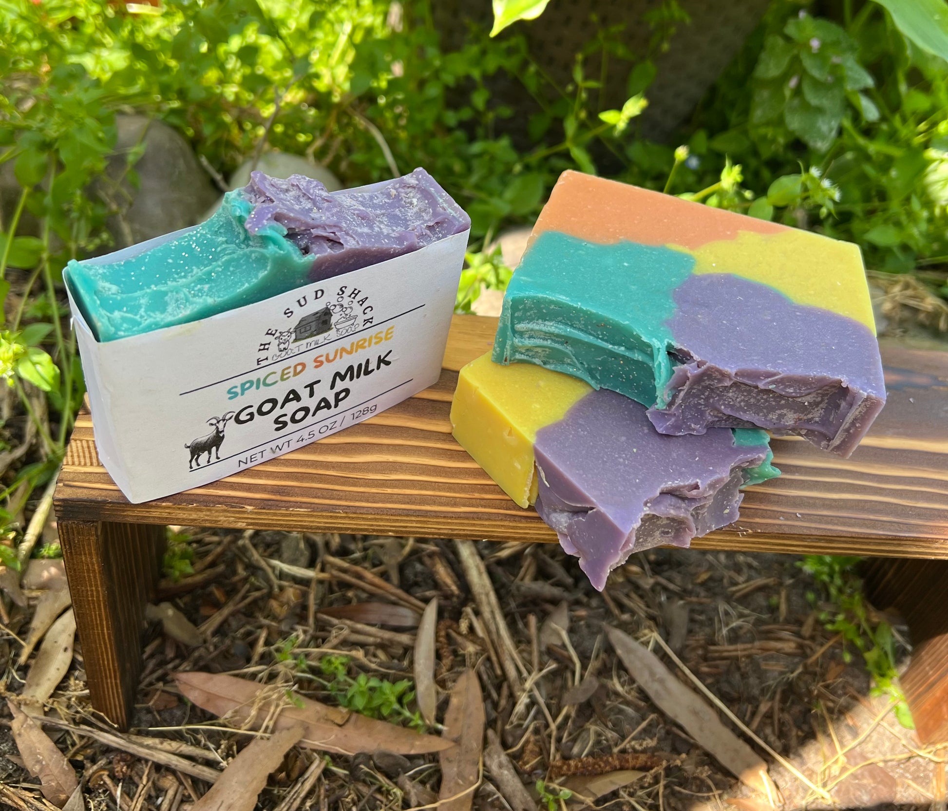 Colorful soap bars on a wooden stand with a box labeled 'Spiced Summer Goat Milk Soap' against a natural background.