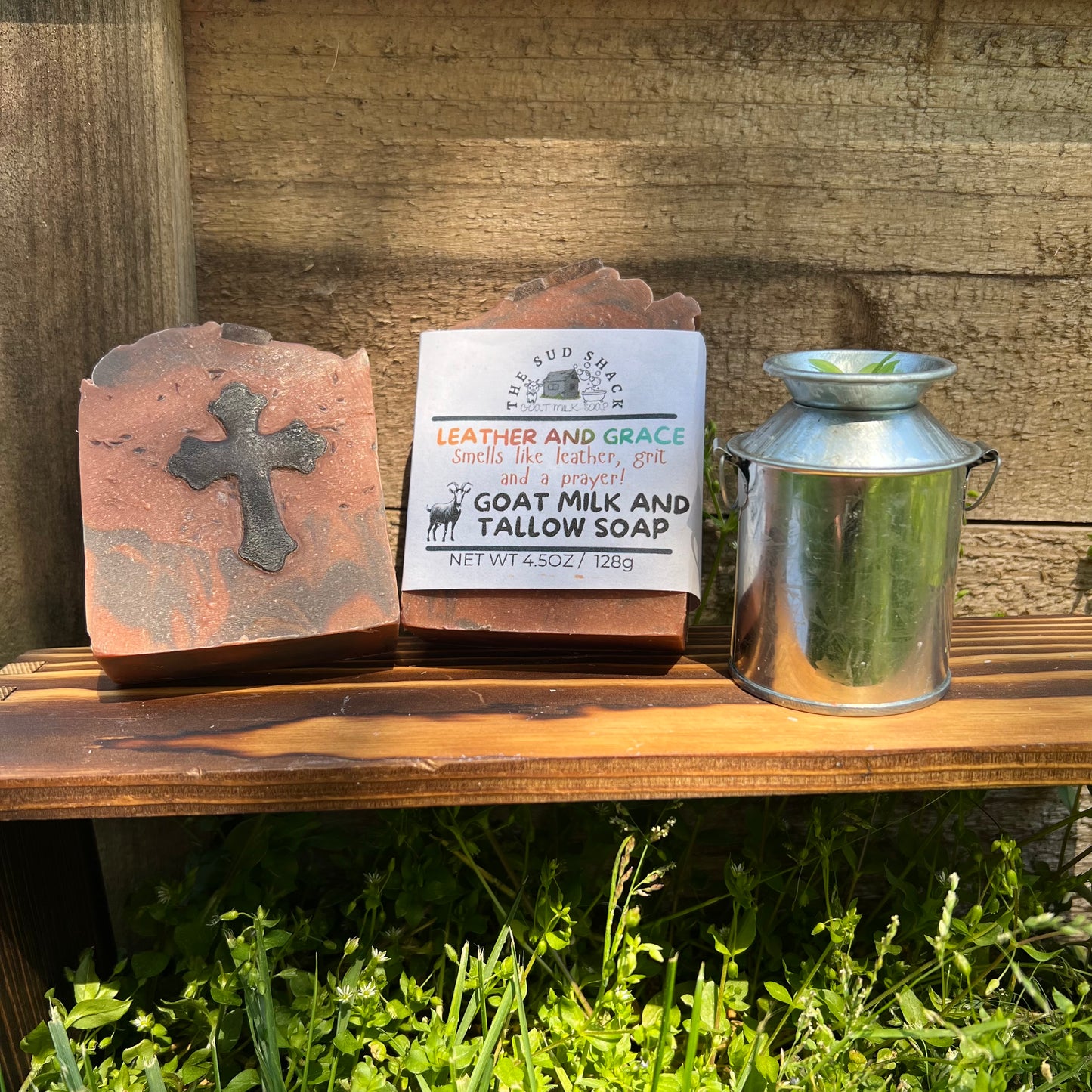 Bar of soap with a cross design, a label, and a metal container on a wooden surface outdoors.