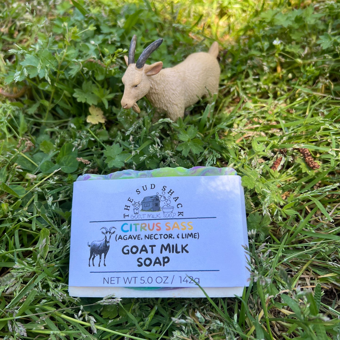 Bar of goat milk soap with a label on grass, accompanied by a small goat figurine.
