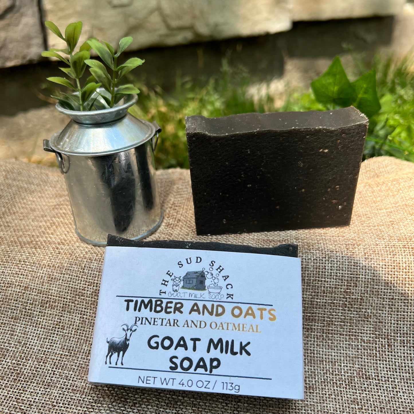 Goat milk soap bar with label on a textured surface, plant in the background