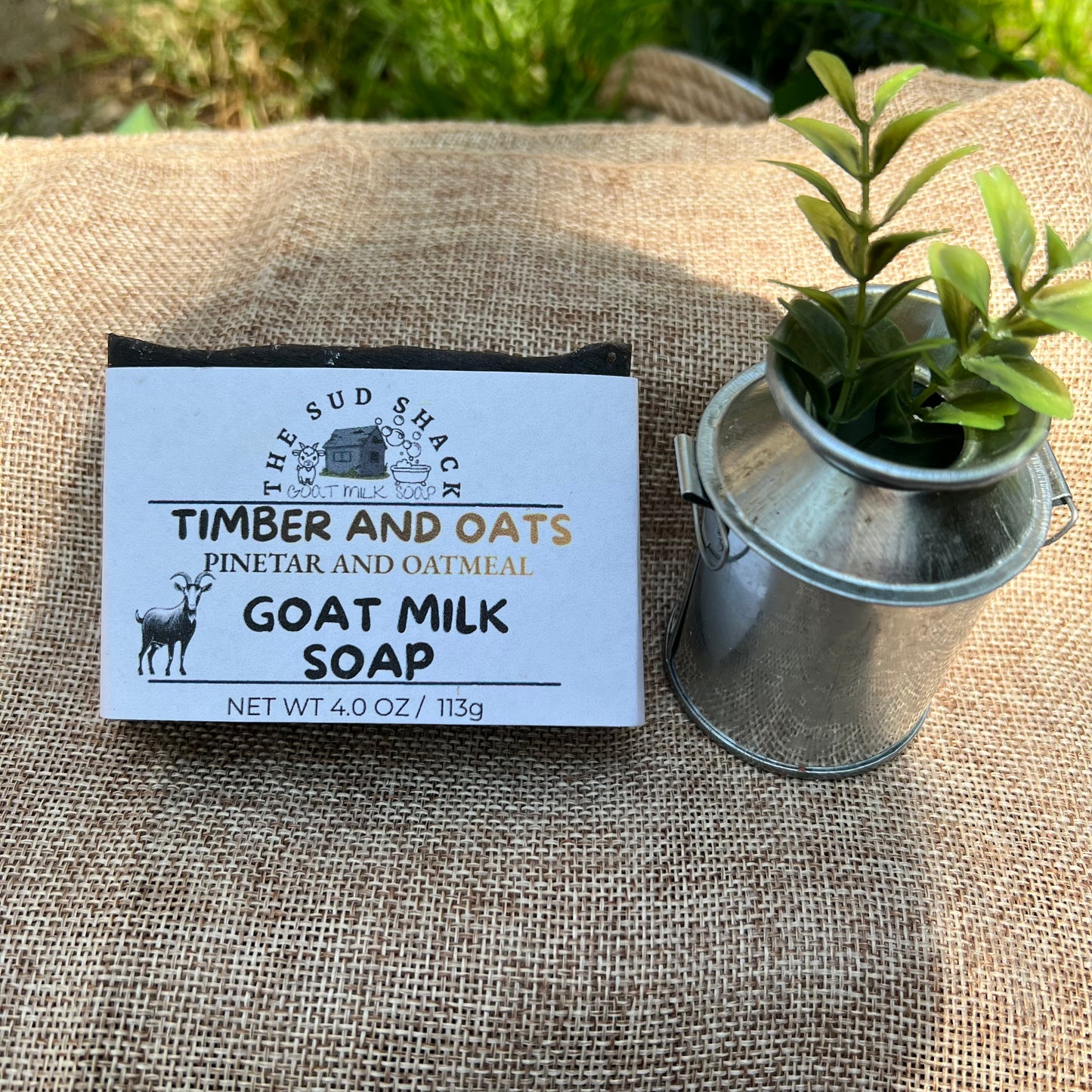 Goat milk soap bar with 'Timber and Oats' label on a textured surface with a plant in the background.