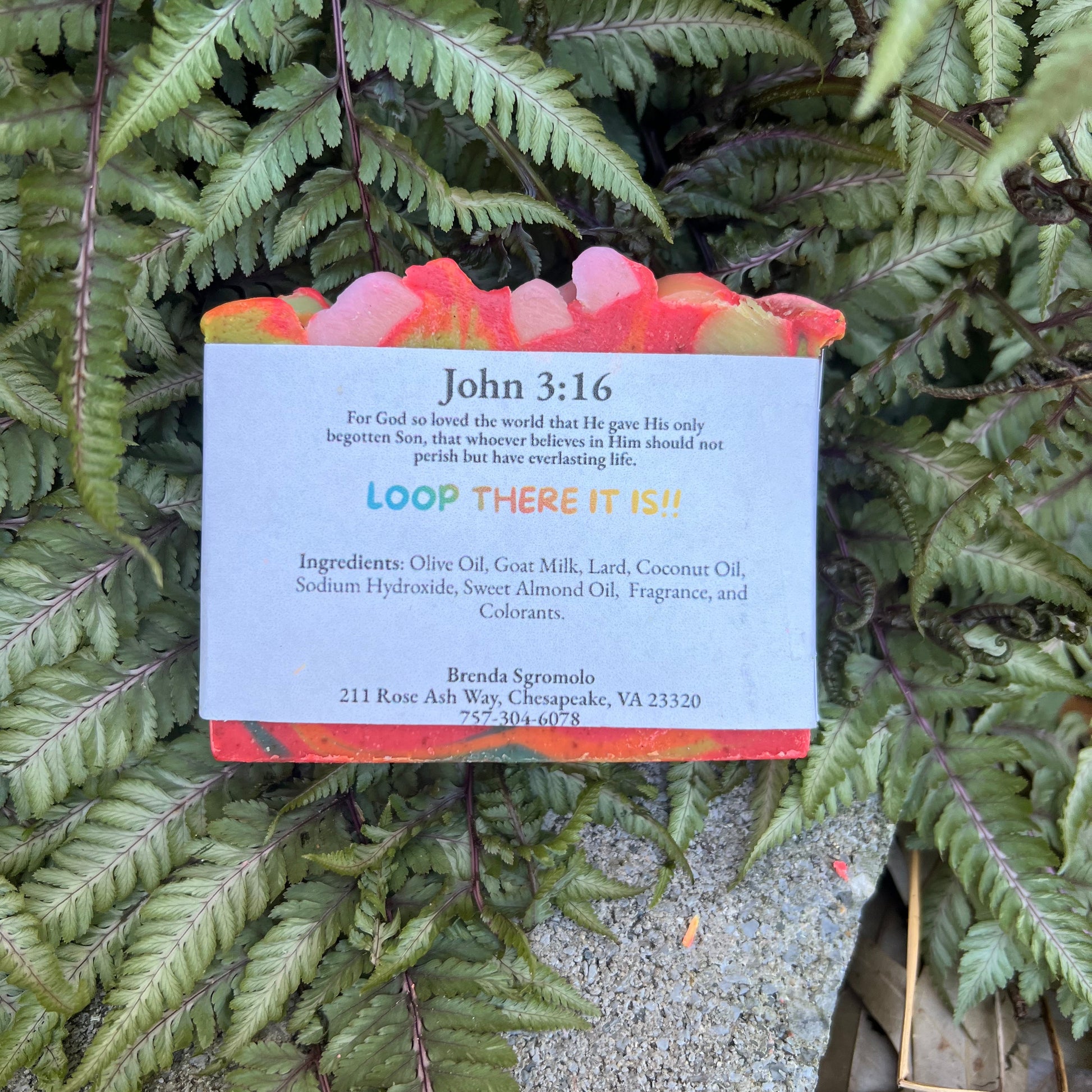 Soap bar with a card displaying 'John 3:16' text and ingredients, placed on green leaves.