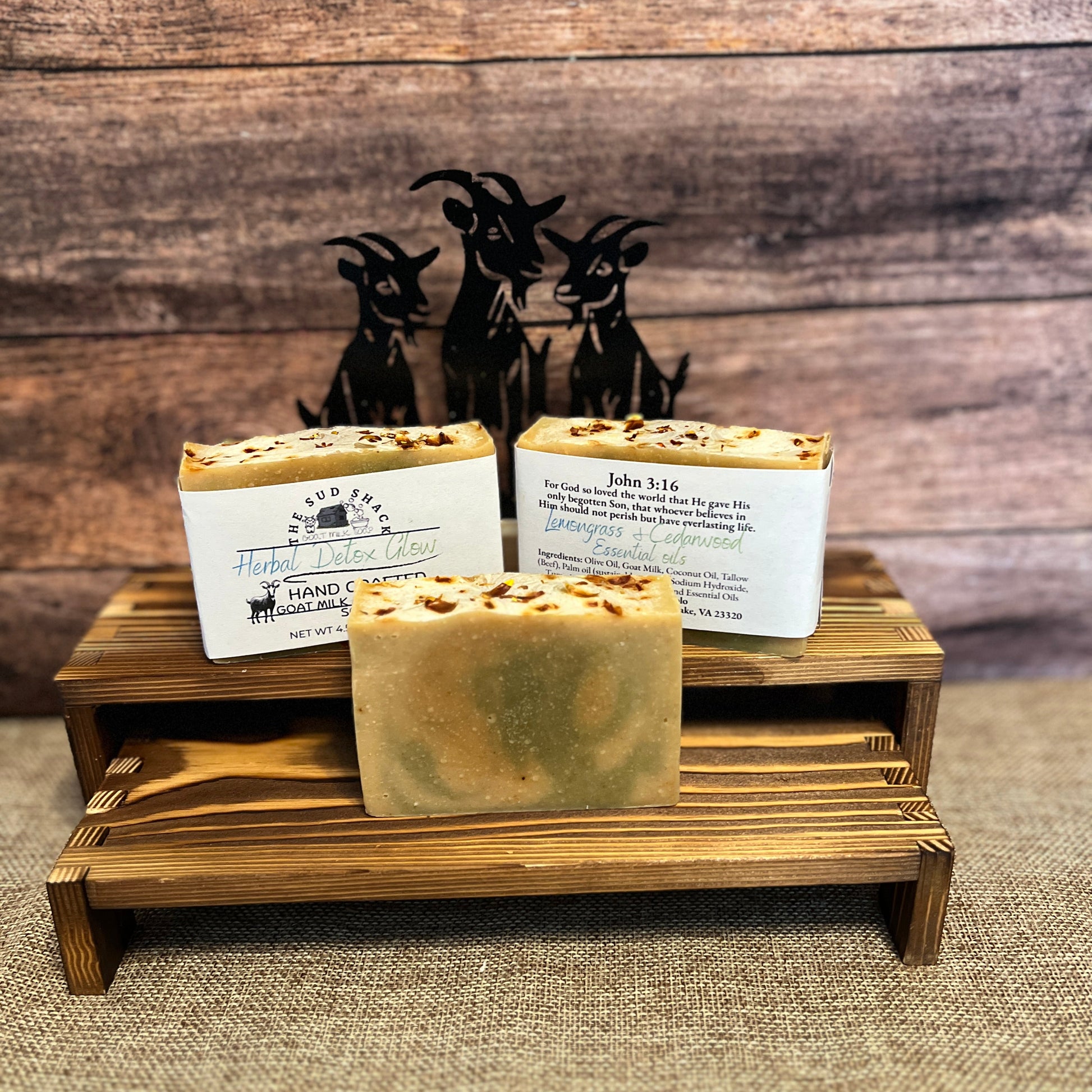 Three bars of soap on a wooden stand with a rustic background featuring metal goat sculptures.